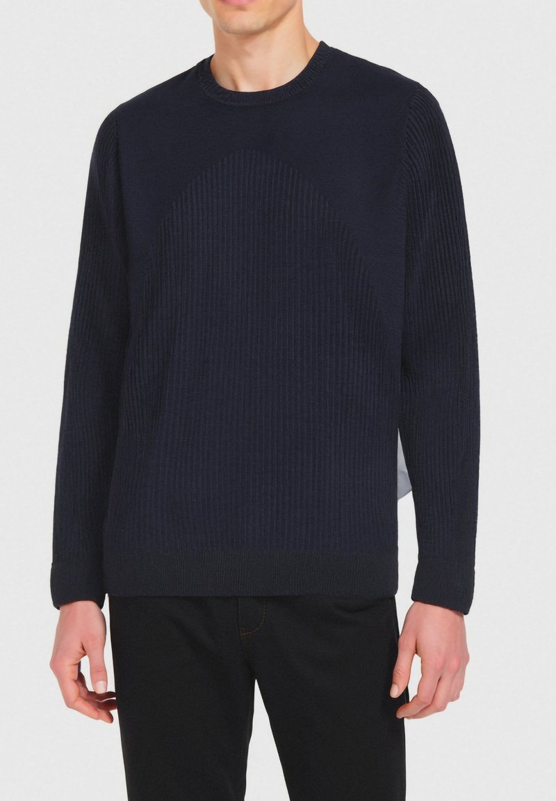 Man in een donkerblauwe geribbelde trui met lange mouwen en zwarte broek, staande tegen een effen lichte achtergrond.