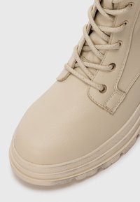 Beige lærstøvle med en glat overflade, udstyret med metaløjer, snørebånd og en tykk sål med et tekstureret dækmønster.