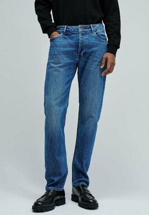 Hombre que lleva jeans de pierna recta azules, camisa negra de manga larga y mocasines de cuero negros, de pie con una mano en el bolsillo.