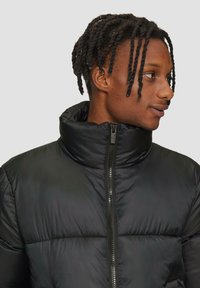 Zwarte pufferjack met een hoge kraag, voorzien van een ritssluiting en een gewatteerde textuur. Glad, lichtgewicht materiaal met een matte afwerking.
