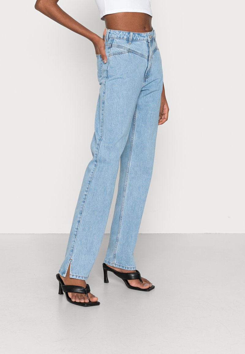 Jeans de cintura alta em azul claro com pernas retas, apresentando uma ligeira fenda na bainha, combinados com sandálias de salto pretas.