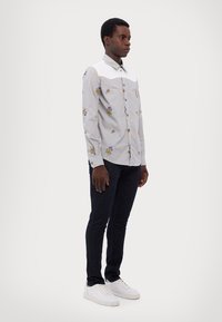 Camicia a maniche corte grigia chiaro con colletto bianco, caratterizzata da ricami floreali blu e gialli, apertura frontale con bottoni e vestibilità slim.