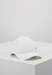 Fehér, ívelt karimájú vászon baseball sapka, elöl egy kicsi fém logóval, a tetején szellőzőlyukakkal.