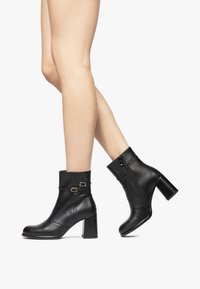 Bottines en cuir noir avec une fermeture éclair, un talon bloc et un revers replié orné de deux boucles décoratives. Texture lisse avec un design épuré.