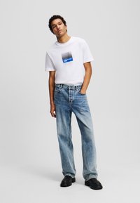 Witte katoenen T-shirt met een blauw en zwart gestreepte vierkante graphic, gecombineerd met lichtblauwe, lose denim jeans en zwarte chunky schoenen.