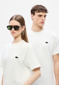 Δύο μοντέλα φορούν λευκά βαμβακερά μπλουζάκια με μαύρο κεντητό λογότυπο στο αριστερό στήθος. Η γυναίκα φοράει μαύρα ορθογώνια γυαλιά ηλίου.