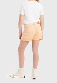 Pantalones cortos de mezclilla en naranja claro con dobladillos enrollados, con bolsillos traseros y una etiqueta de cuero marrón en la cintura. Combinados con zapatillas blancas.