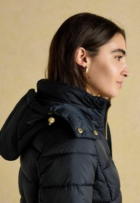 Žena v čiernej páperovej bunde s kapucňou a zlatým zipsom, otočená doprava pred béžovým pozadím.