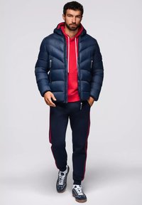 Giacca puffer blu navy con cappuccio sopra un hoodie rosso, abbinata a pantaloni della tuta blu navy con strisce laterali rosse e sneakers blu navy con dettagli bianchi.