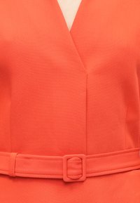 Koralloranž värvi kleit, millel on V-kaelus, struktureeritud kangas ja vöökohal ruudukujulise pandlaga sobiv vöö.