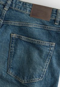 Keskitummansiniset farkut, joissa on teksturoitu pinta, takatasku ja ruskea nahkalappu, johon on painettu "DENIM SUPPLY".