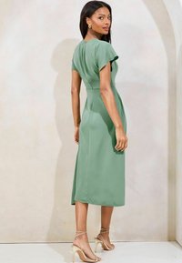 Vestito midi verde con maniche corte a sbuffo, vita aderente e gonna fluida. Indossato con tacchi nudi con cinturini. Tessuto liscio.