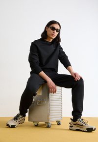 Felpa nera e pantaloni in tessuto morbido. Il modello è seduto su un contenitore metallico. Indossa occhiali da sole neri e scarpe da ginnastica beige.