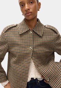 Bruine en zwarte houndstooth jas met een grote kraag, zilveren knopen en gestructureerde schouders, gedragen over een geplooide witte blouse.