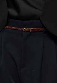 Πλεκτή καφέ δερμάτινη ζώνη με χρυσή αγκράφα, στερεωμένη γύρω από μαύρο παντελόνι κομμένο στα μέτρα με πουκάμισο που έχει φορεθεί μέσα.