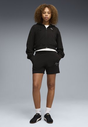 Ung kvinde med krøllet hår står, iført sort Puma lynlås-hættetrøje, matchende shorts, hvide sokker og sorte Puma sneakers.