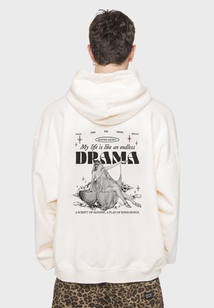 Sudadera color crema con un estampado gráfico en la parte posterior que presenta una figura sentada, texto y elementos decorativos. Tejido suave, corte relajado.