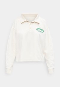 Εκρού πόλο μπλούζα με μακριά μανίκια και γιακά, με πράσινο κείμενο "Calvin Klein New York City" στο αριστερό στήθος.