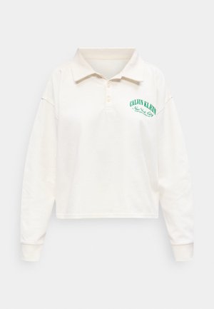 Εκρού πόλο μπλούζα με μακριά μανίκια και γιακά, με πράσινο κείμενο "Calvin Klein New York City" στο αριστερό στήθος.