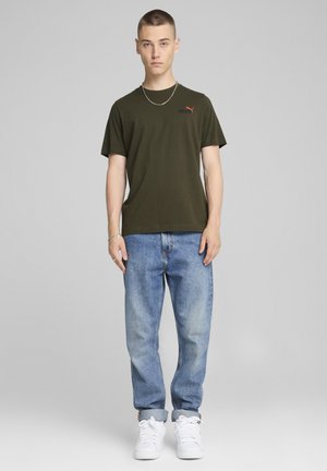 Camiseta de algodón verde oliva con mangas cortas, pequeño logotipo rojo y negro, combinada con jeans de mezclilla azul claro y zapatillas blancas.