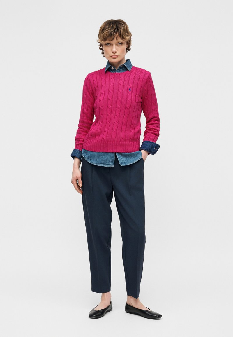 Personne portant un pull en maille torsadée rose vif par-dessus une chemise en denim, un pantalon plissé bleu marine et des ballerines noires, debout devant un fond uni.