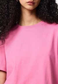 Jeune femme aux cheveux bouclés et foncés portant un t-shirt rose uni à col rond et une petite boucle d'oreille, lèvres légèrement entrouvertes.
