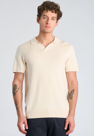 Jonge man met krullend haar, gekleed in een crème poloshirt met korte mouwen en donkere broek, toont tatoeages op beide onderarmen, staand tegen een lichte achtergrond.
