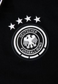 Černá látka s bílým kruhovým logem Deutscher Fussball-Bund, které zobrazuje orla a čtyři vyšívané hvězdy nad ním.