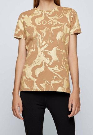 Lyhythihainen t-paita, jossa on kultaiset abstraktit kieputukset ja "BOSS"-teksti, yhdistettynä mustiin istuviin housuihin, edestä kuvattuna olkapäiltä lantioon.