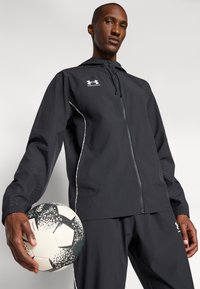 Jachetă neagră cu fermoar, glugă și accente laterale reflective, purtată cu pantaloni negri asortați; ținând în mână o minge de fotbal albă cu un model negru.