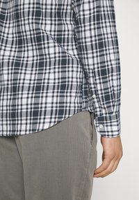 Gros plan sur le torse d'une personne portant une chemise à manches longues à carreaux noirs et blancs avec un pantalon beige, la main détendue le long du corps.