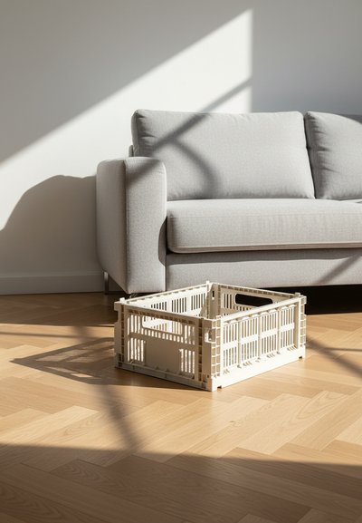 Caisse en plastique beige sur un sol en bois à côté d'un canapé gris clair avec la lumière du soleil et les ombres projetées sur les deux.