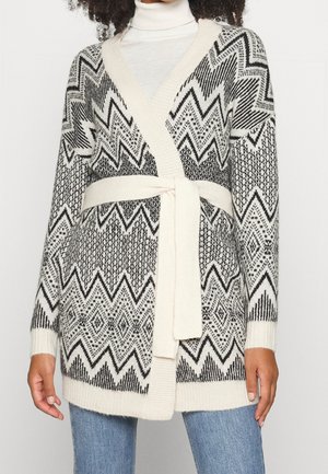 Femme portant un cardigan en maille noir et blanc avec des motifs géométriques et une ceinture nouée, sur un col roulé blanc et un jean bleu.