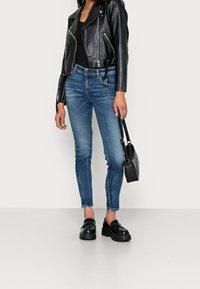 Chaqueta de moto de cuero negro, camiseta negra ajustada, vaqueros ajustados azules, mocasines negros gruesos y un pequeño bolso negro.
