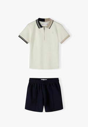 Krém rövid ujjú póló fekete és bézs csíkos gallérral és mandzsettákkal, fekete elasztikus derékpántos rövidnadrággal párosítva.