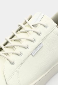 Sneaker en cuir blanc avec surface texturée, bout rond, lacets plats et une étiquette de marque sur le côté. Doublure intérieure lisse.