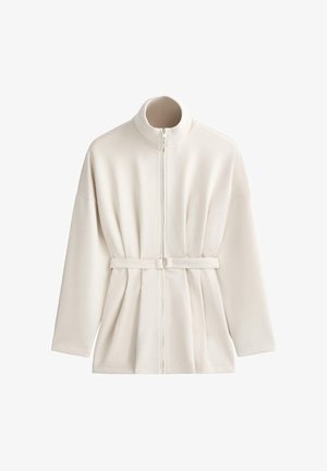 Dlhá bunda krémovej farby s vysokým golierom, predným zipsom a nastaviteľným opaskom v páse, zobrazená na bielom pozadí.