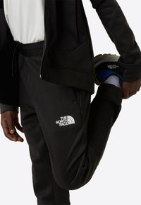 Svarte joggebukser med hvit logo på låret, med snøring i midjen og teksturert detaljering på nedre del av benet.