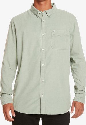 Homme portant une chemise boutonnée vert clair à manches longues avec une poche poitrine et des boutons blancs, debout devant un fond uni.