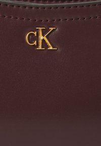 Cerca de un cuero burdeos oscuro con una textura suave. Presenta un logo dorado "cK" destacado. Detalles de costura a lo largo de los bordes.