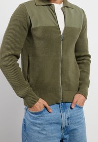 Olivgrön zip-up cardigan med ribbad textur, kontrasterande släta paneler och sidfickor. Fitted silhuett med krage.