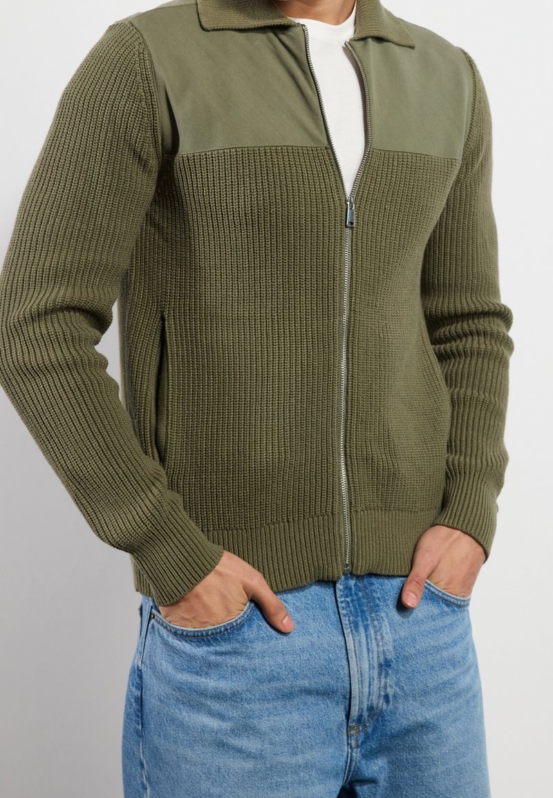Olivgrön zip-up cardigan med ribbad textur, kontrasterande släta paneler och sidfickor. Fitted silhuett med krage.