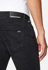 Jeans de mezclilla negra con una textura suave, que presentan una etiqueta MAVI en la cintura y un diseño sutil en el bolsillo trasero.