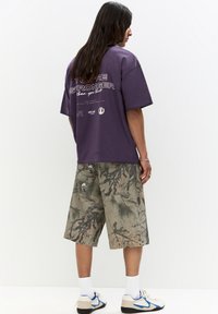 Fialové oversized tričko s bílým textem na zádech, spárované s šortkami s maskáčovým vzorem. Model má na sobě bílo-modré tenisky.