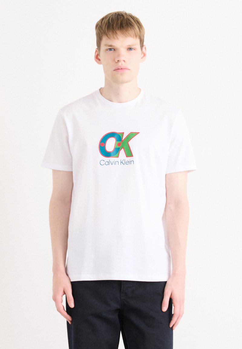 White cotton t-shirt featuring a large, colorful "OK" logo and "Calvin Klein" text. Simple crew neck and short sleeves.