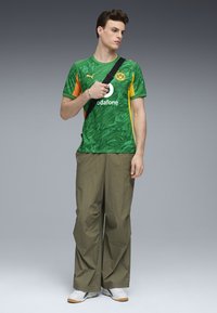 Jersey verde con mármol y acentos naranjas, correa cruzada negra, pantalones olivo holgados y zapatillas blancas con suelas beige.