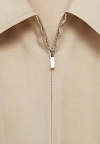 Camicia beige con zip frontale, colletto a punta e texture in tessuto liscia. Presenta una zip argentata con un tirante rettangolare.