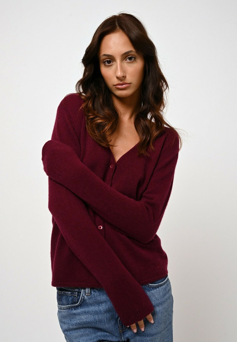 Cardigan bordeaux con scollo a V, maniche lunghe e chiusura a bottoni. Texture morbida con dettagli a coste sui polsini, abbinato a jeans di denim chiaro.