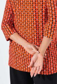 Punainen ja oranssi kuvioitu paita, jossa on geometrinen muotoilu, 3/4-hihat ja pyöreä pääntie. Mustat housut; kädet ristissä selän takana.