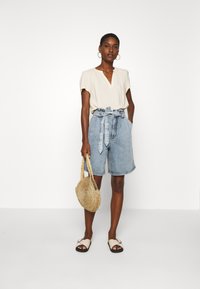 Ljus beige t-shirt med kort ärm och v-ringning, i kombination med blå denim shorts med hög midja och platta sandaler. En vävd stråhandväska ingår.
